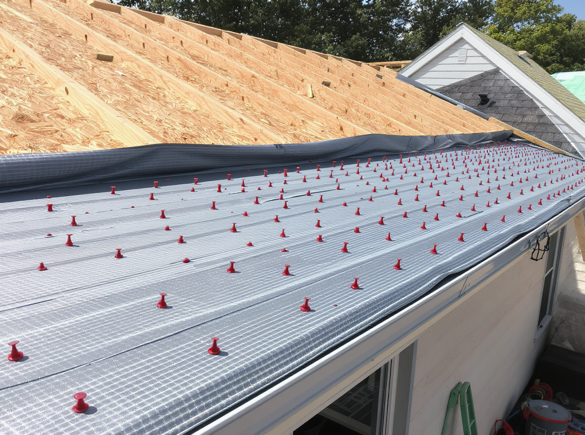 Synthetic underlayment being rolled across plywood roof decking during a residential roof replacement in Baldwin County Alabama with red plastic cap nails securing the weather barrier and galvanized metal drip edge installed at the eave line, a critical step before architectural shingle installation that protects against wind-driven rain infiltration in Gulf Coast conditions with 66 inches of annual rainfall